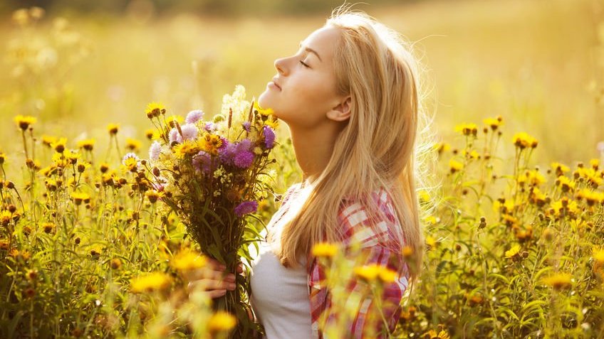 Mulher de perfil segurando buquê de flores em campo com flores amarelas iluminada pelo sol