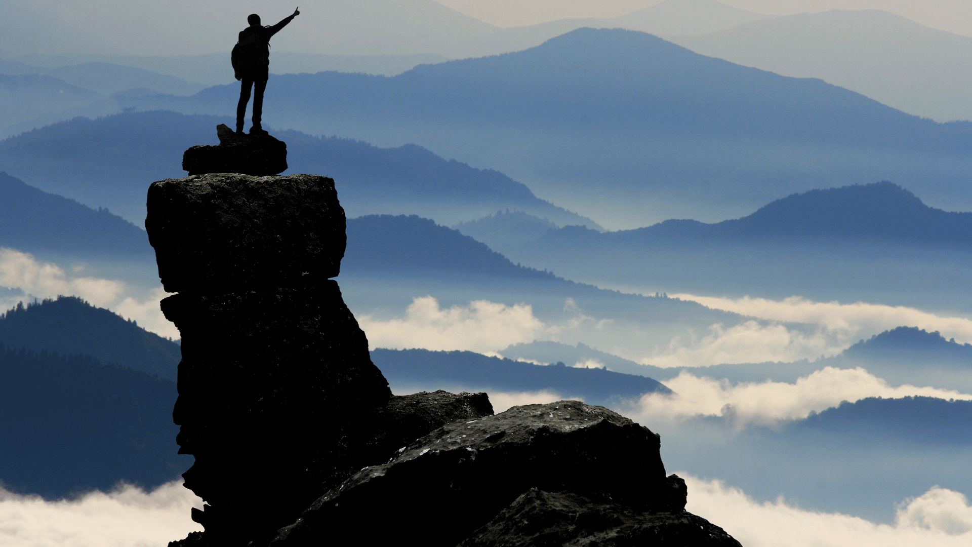 Homem de pé sobre uma montanha de pedras. Ele está com uma mochila nas costas. Ao fundo montanhas e nuvens de tons azuis e branco.