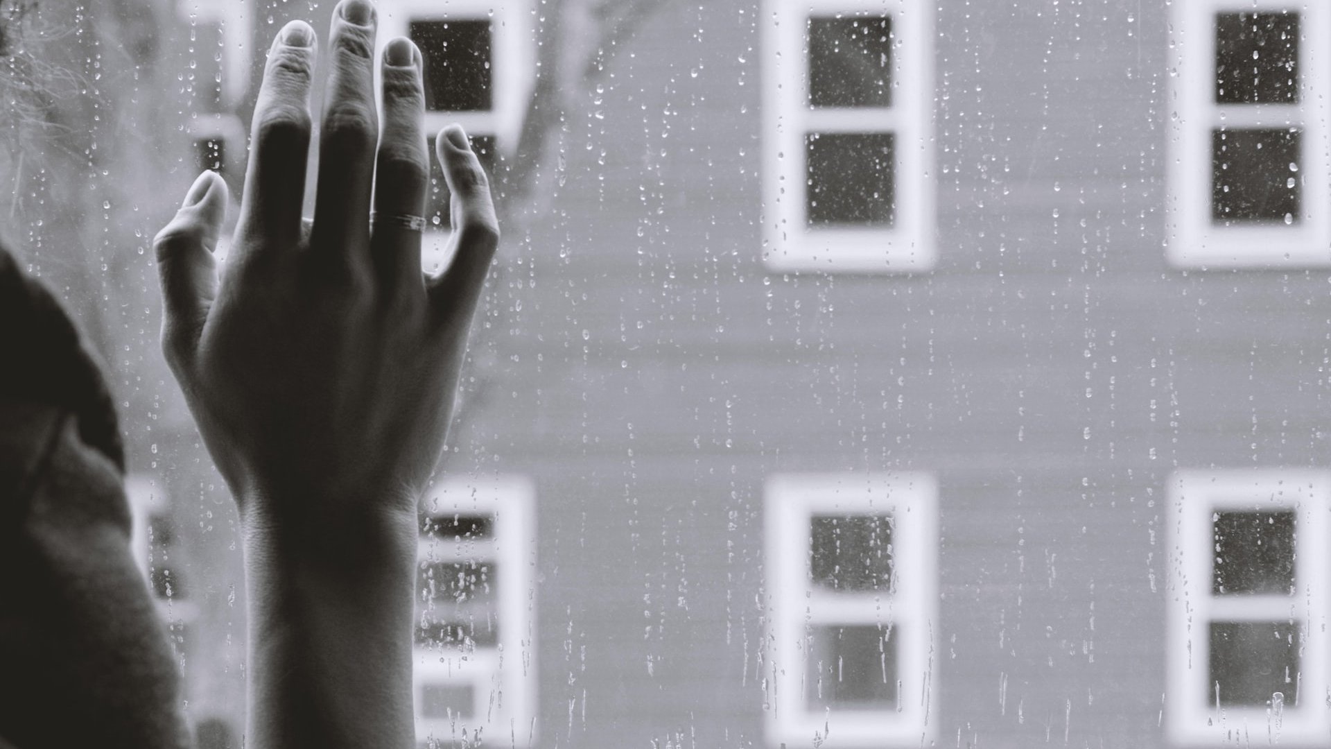 Mão feminina tocando uma janela com gotas de chuva escorrendo por ela.