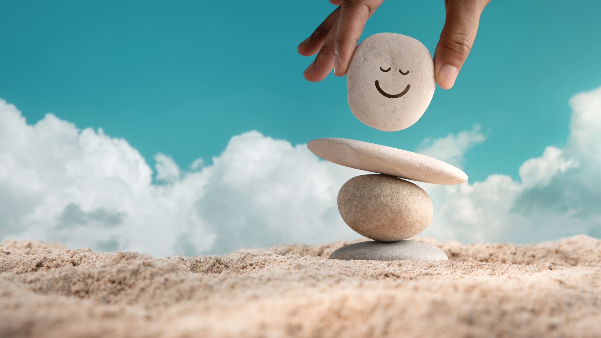 Imagem de um céu azul com algumas nuvens brancas ao fundo. Um piso de areia e sobre ele, algumas pedras sendo seguradas pelas mãos de uma pessoa, compõe a imagem. A última pedra, traz o sorriso de uma pessoa.