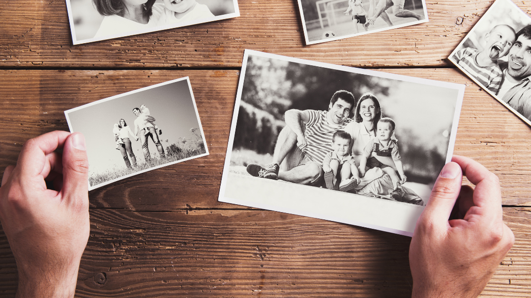 Uma mão está segurando fotos reveladas, em preto e branco, da infância e da família.