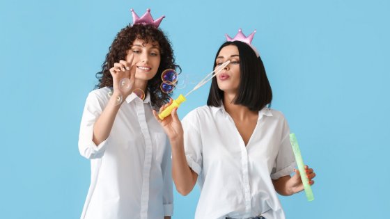 Duas mulheres com coroas de brinquedo em suas cabeças, uma fazendo bola de sabão e a outra as estourando.