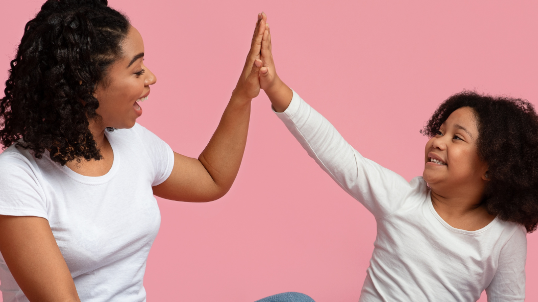 Mãe e filha sentadas no chão dando um high five (levantando a mão e batendo na mão da outra).