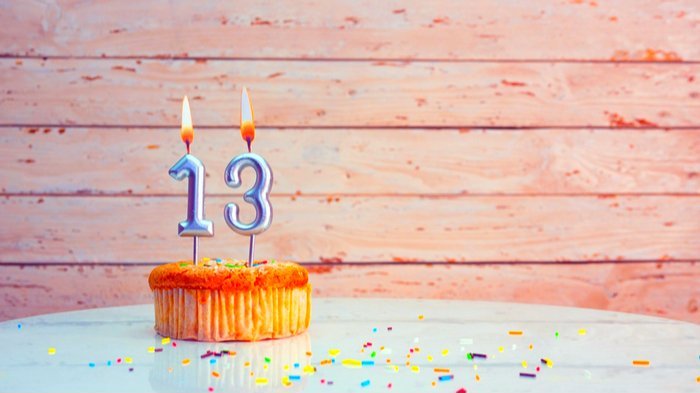 Bolo de aniversário bem pequeno com velas em formato do número 13 acesas em seu topo. Na mesa sob ele há confetes coloridos espalhados.