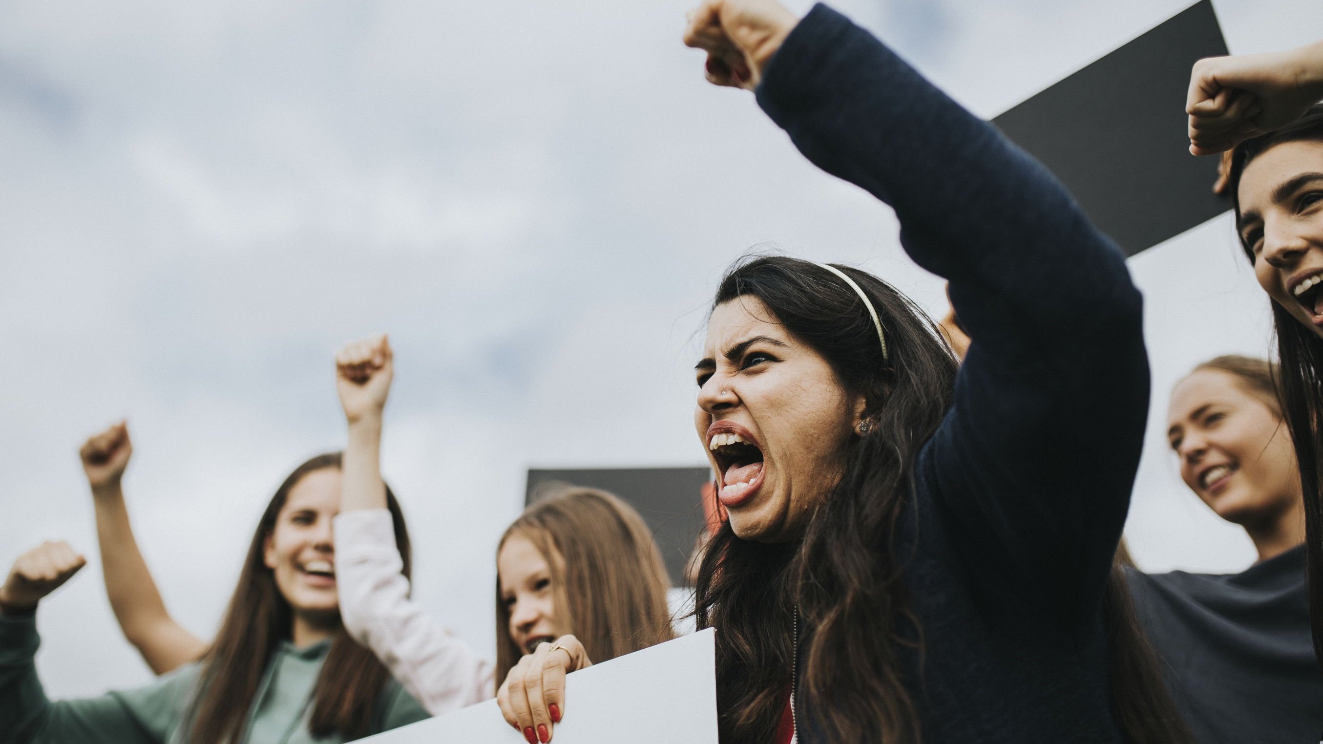 Mulheres protestando segurando cartazes e ergendo seus punhos para o alto.