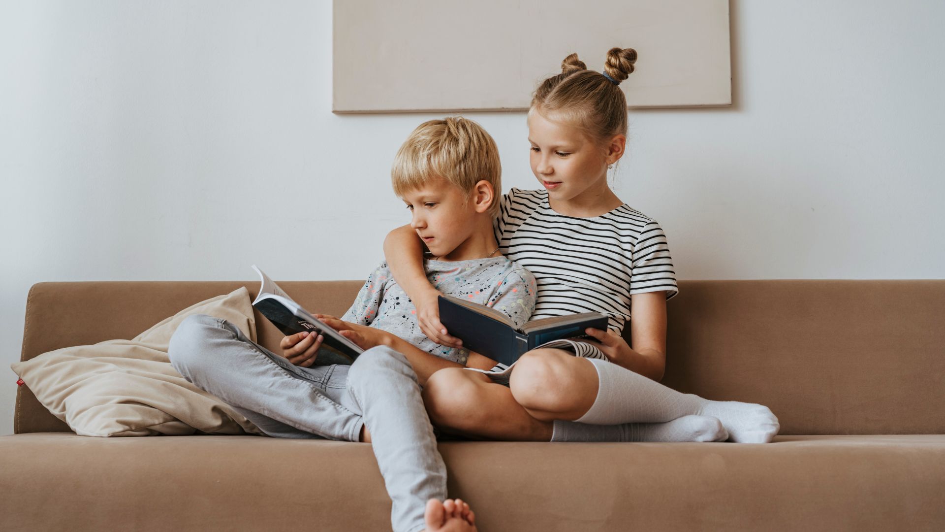 Imagem de uma irmã e um irmão sentados no sofá e lendo livros.