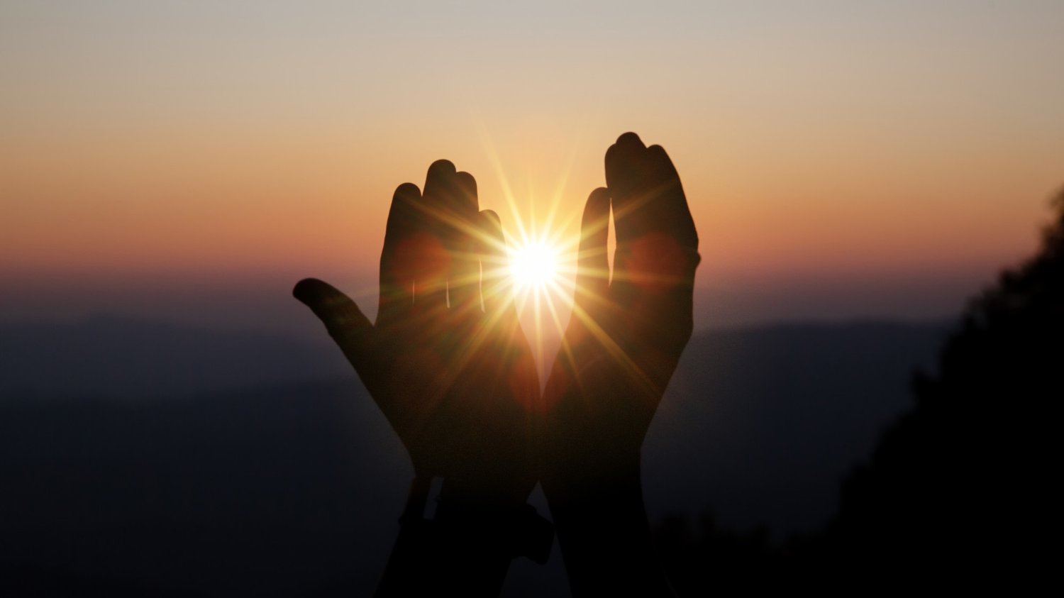 Mãos entreabertas mostrando o sol, ao fundo, o pôr do sol