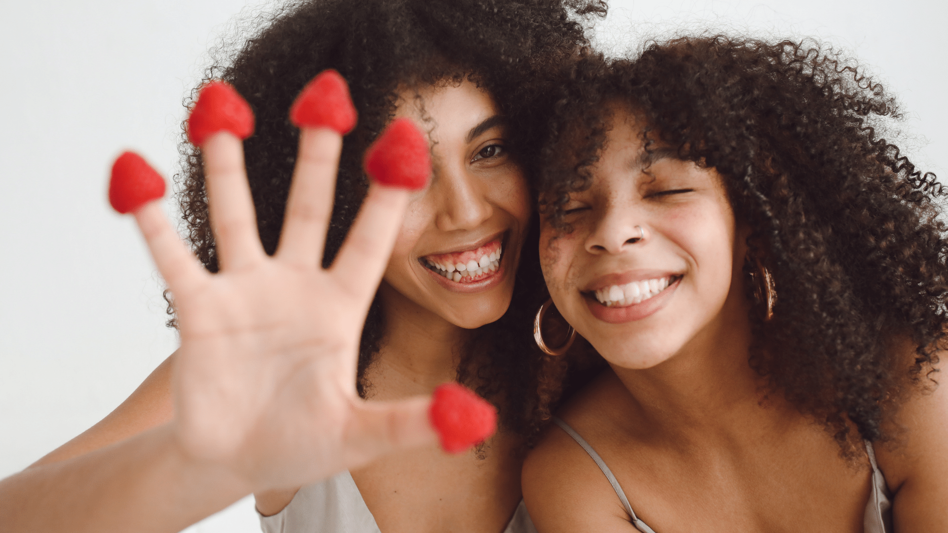 Foto de duas irmãs sorrindo, uma está com as mãos para frente e tem morangos nas pontas de seus dedos.