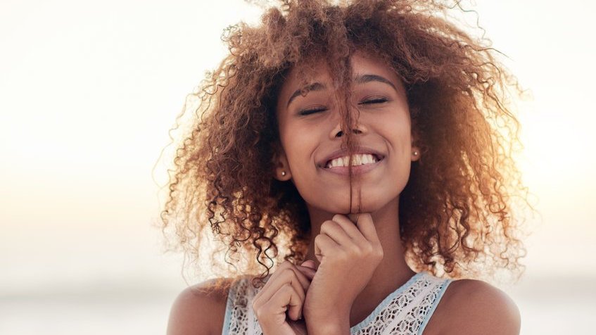 Mulher negra com os olhos fechados, sorrindo e segurando cabelo.