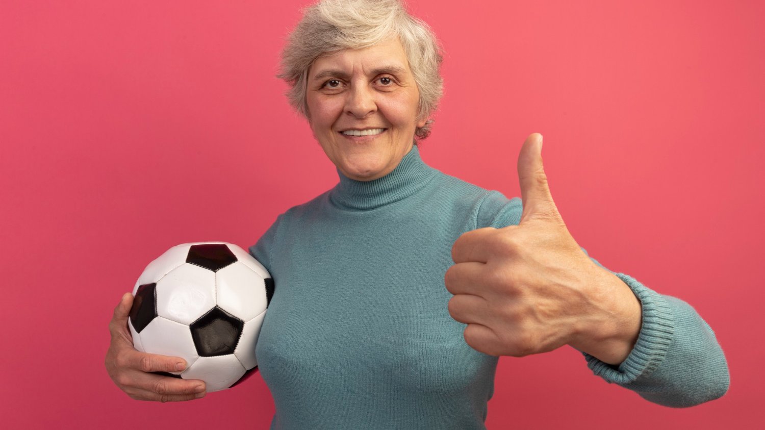 Mulher idosa segurando uma bola de futebol e fazendo joia com a mão