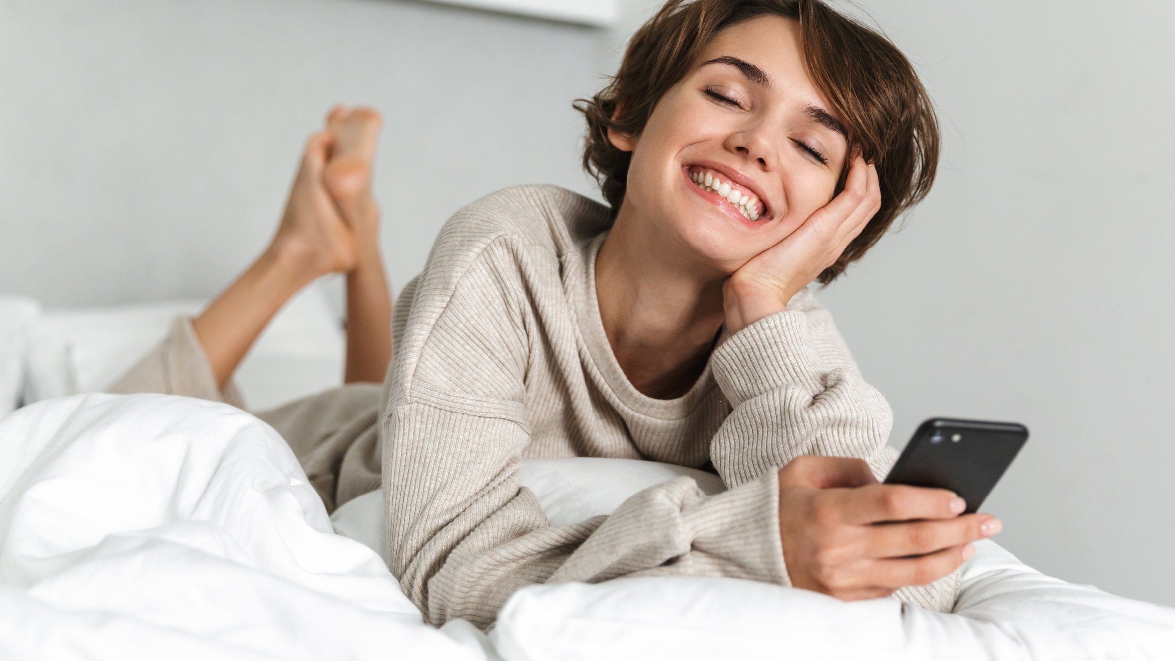 Mulher branca deitada numa cama, segurando celular e com expressão sorridente.