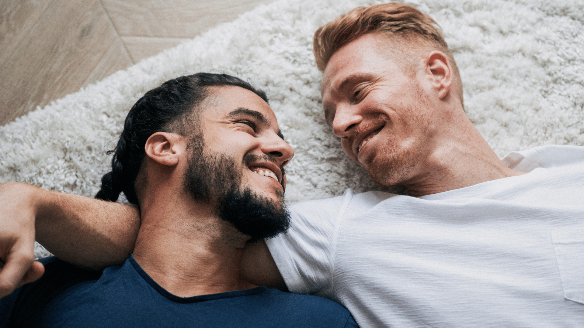 Foto de casal gay, estão olhando um para o outro e sorrindo e deitados no tapete.