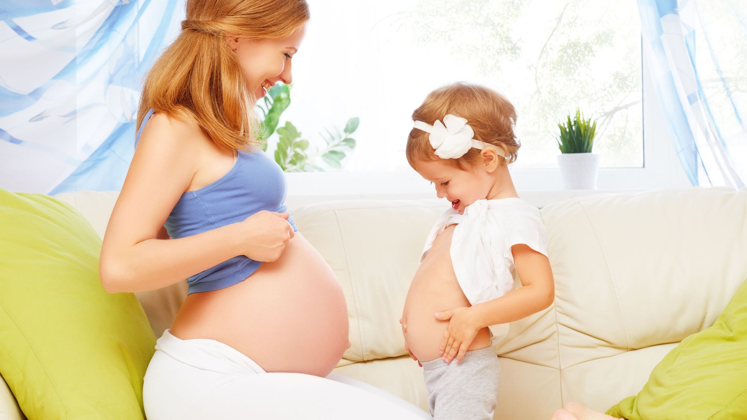 Mulher com blusa que deixa sua barriga grávida a mostra, sorrindo, de frente para uma menina pequena, que segura sua blusa levantada enquanto estufa a barriga para parecer grande.