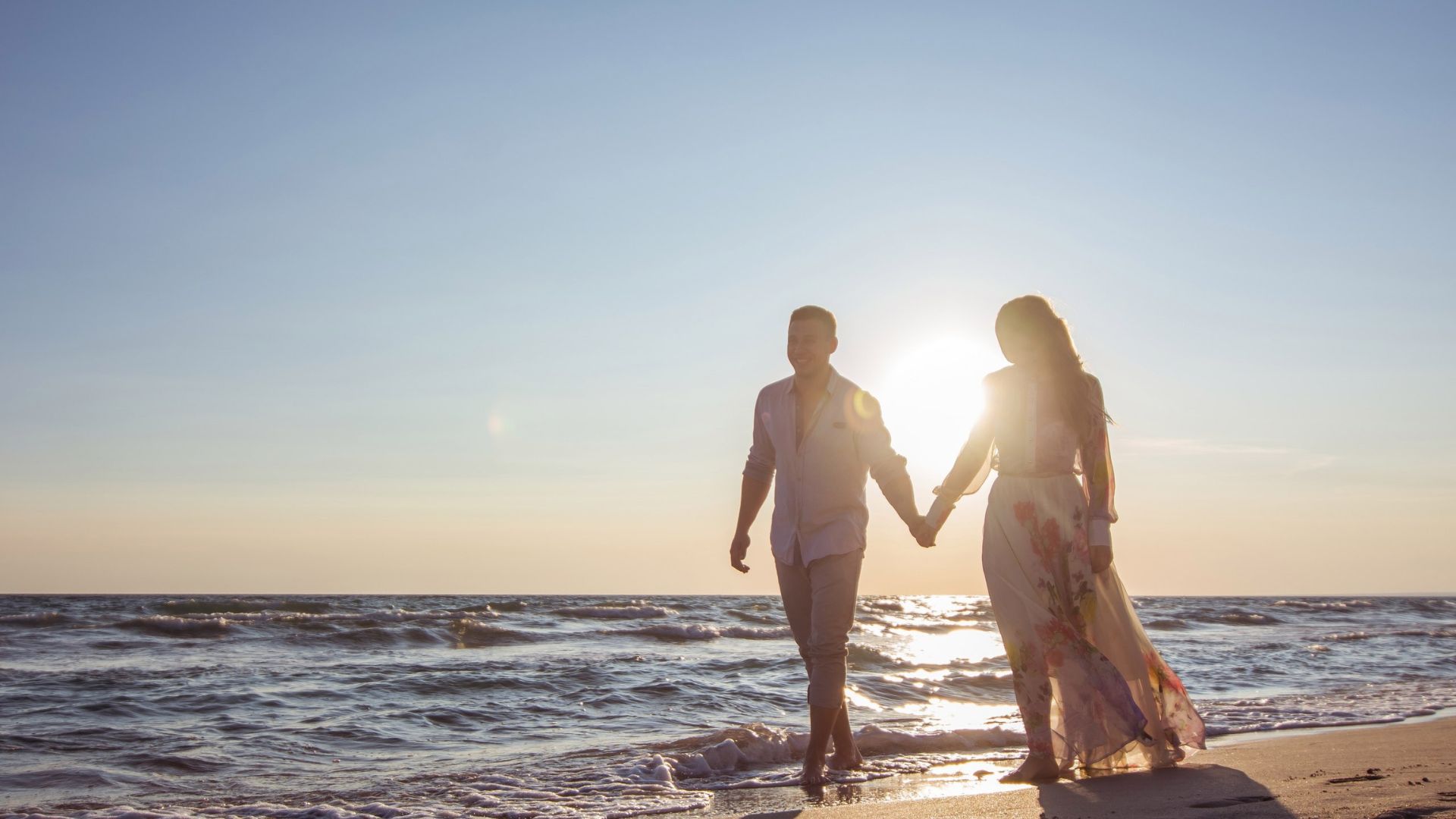 Imagem do mar e ao lado, temos um casal de mãos dadas caminhando na areia.