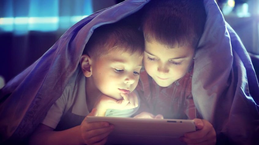 Dois irmãos embaixo de uma coberta mexendo no tablet