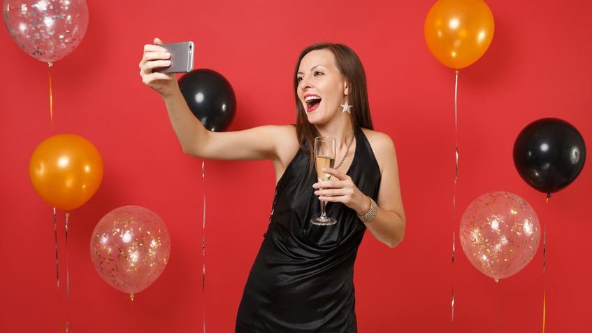 Mulher jovem em um vestido preto, segurando a taça de champanhe, fazendo selfie com balões de ar de fundo vermelho brilhante.
