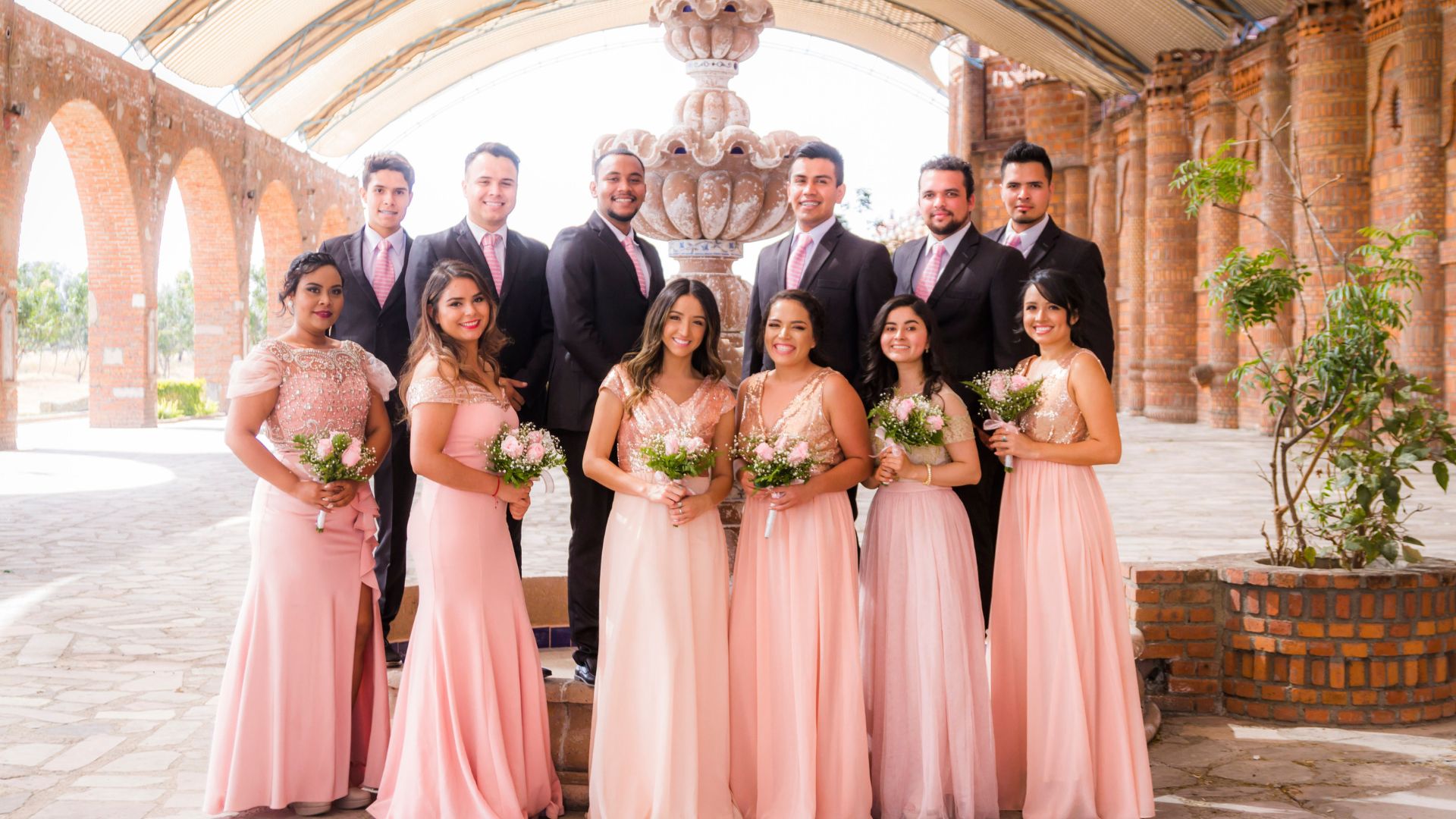 Imagem de uma foto de padrinhos e madrinhas de casamento. Os homens usam terno escuro e gravata na cor rosa e as mulheres, vestido longo na cor rosa claro. Todas seguram um buquê de rosas na mesma tonalidade do vestido.