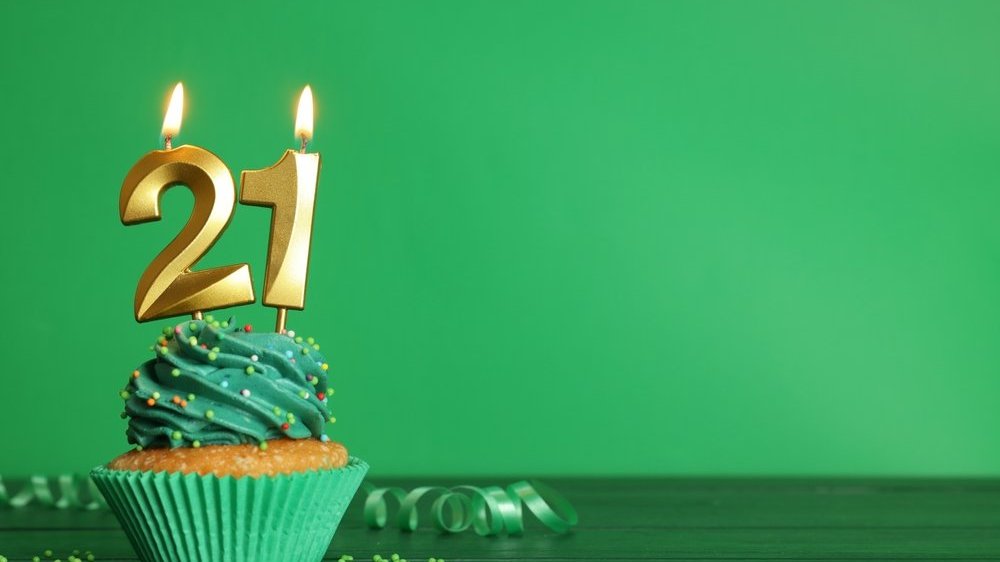 Cupcake com forma e cobertura verdes, e duas velas acesas formando o número vinte e um espetadas no topo. A mesa e a parede ao fundo também são em tons de verde.