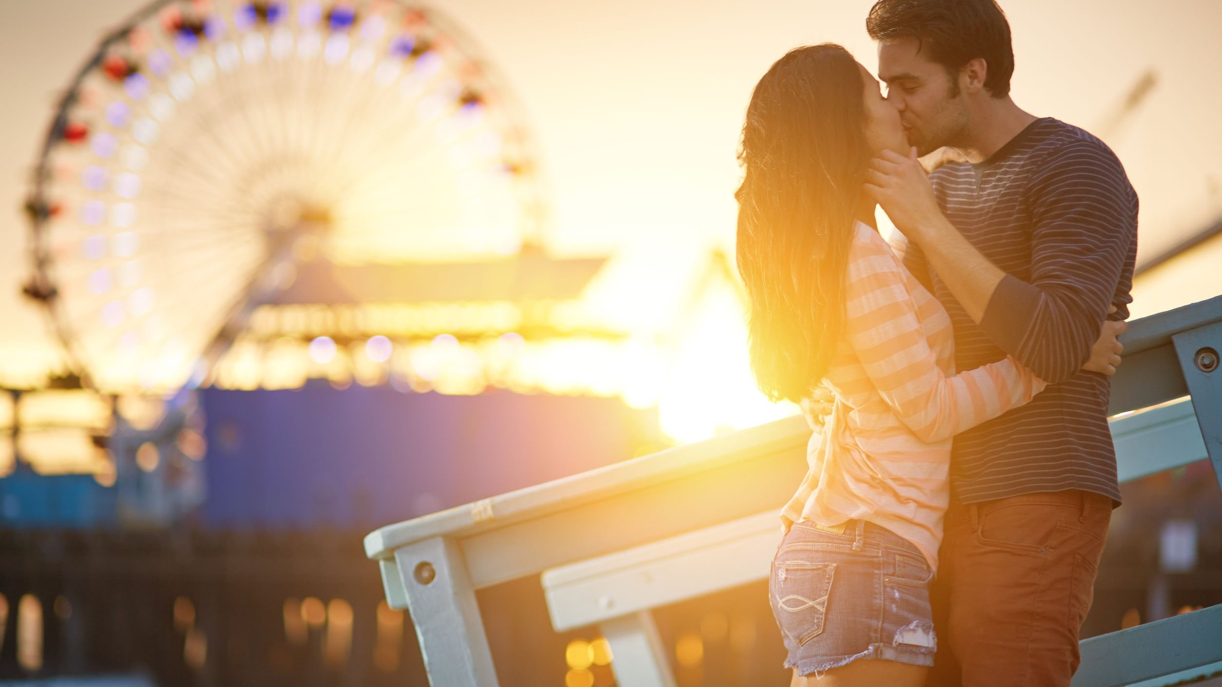 Homem e mulher se beijando com o pôr do sol e uma roda-gigante no fundo. 123rf - Joshua Resnick