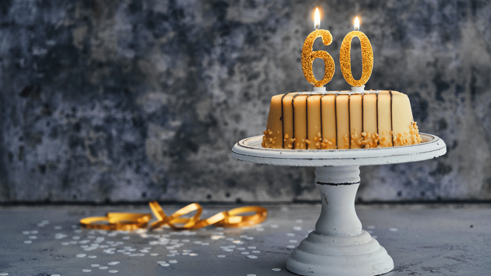 Imagem de um bolo dourado e uma velinha de 60 anos no topo, em cima de uma mesa com confetes dourados.