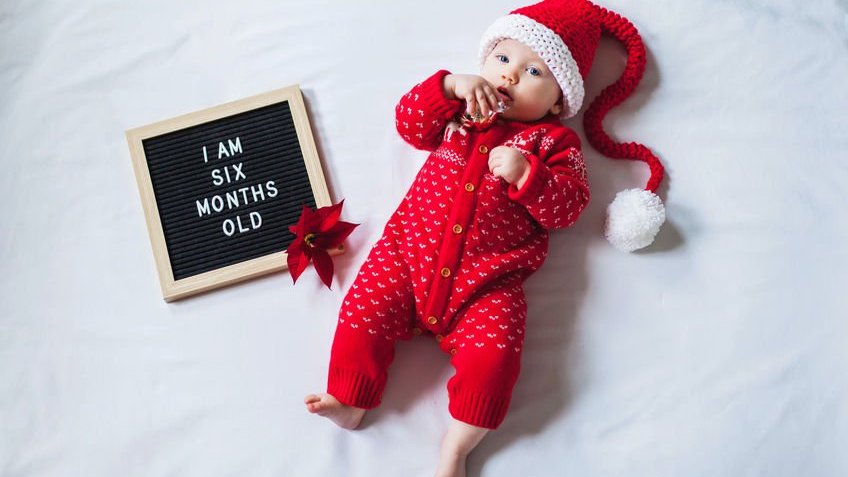 Bebê com roupas natalinas e placa ao lado com os dizeres tenho seis meses de idade