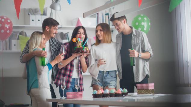 Amigos comemorando aniversário. Uma menina está segurando o bolo para assoprar as velas