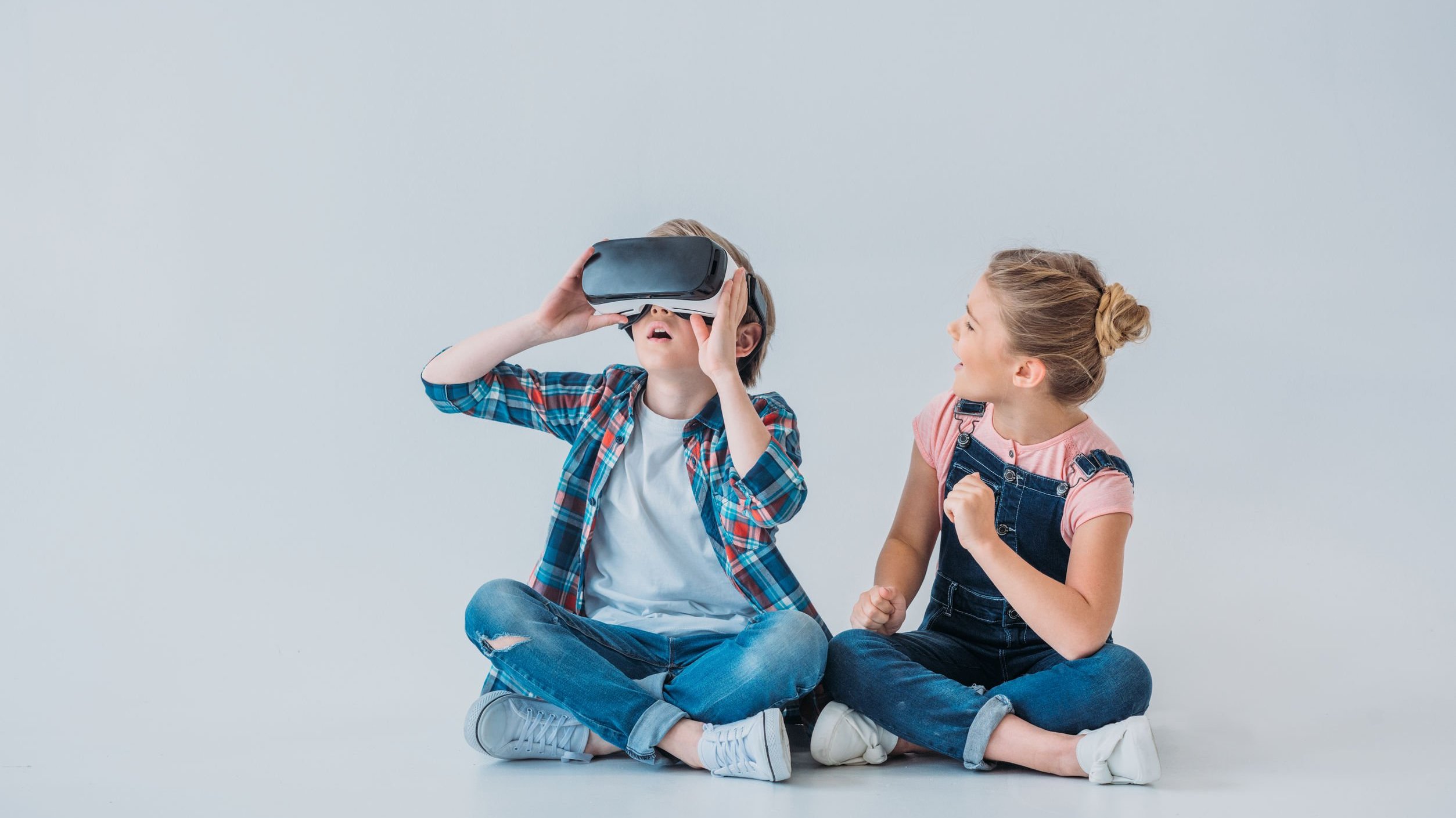 Menino e menina brancos sentados no chão brincando, o menino com um óculos de realidade virtual