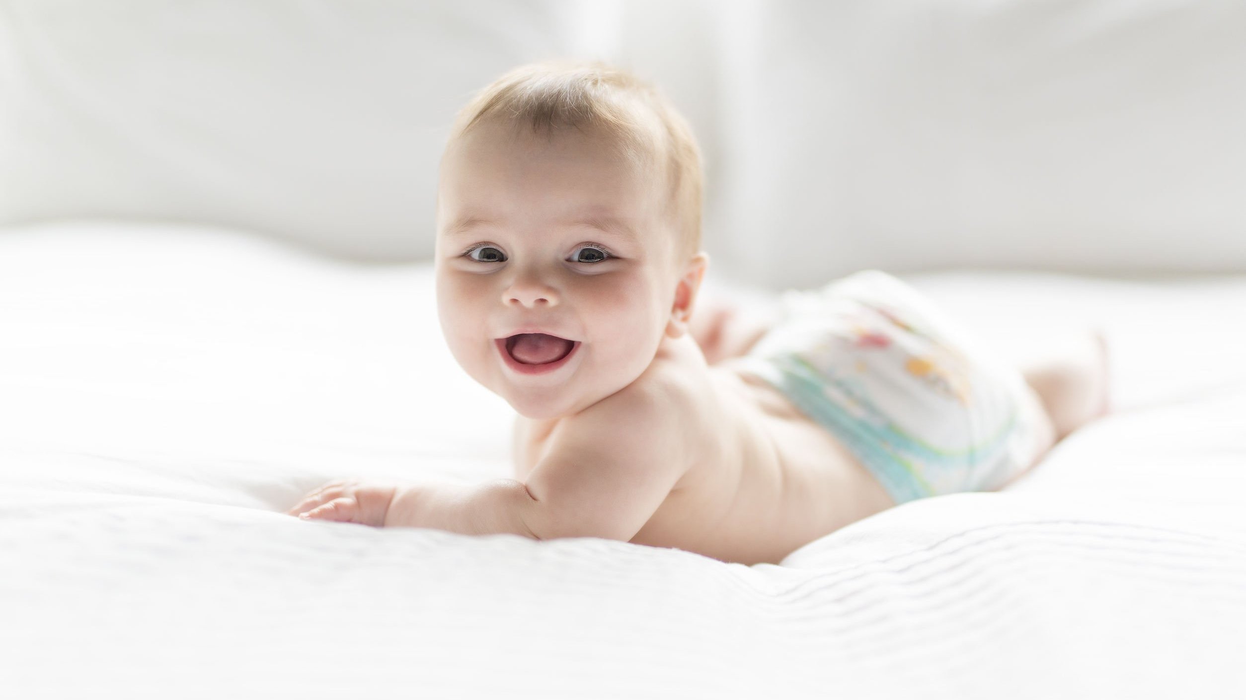 Bebê de 7 meses usando fraldas, deitado de bruços em uma cama enquanto sorri.