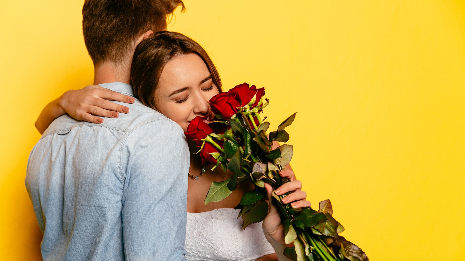 Casal se abraçando enquanto mulher sorri e cheira buquê de rosas