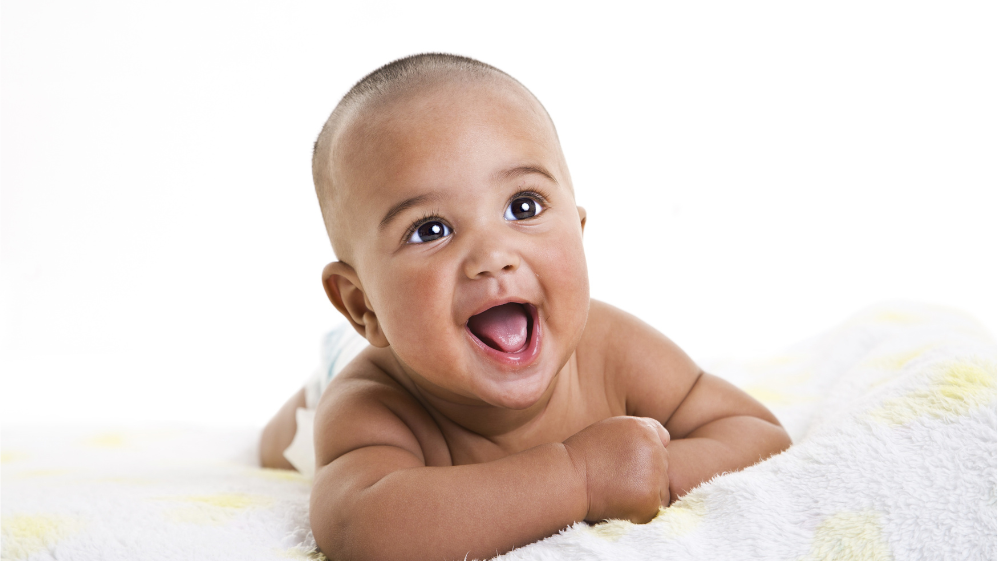 Bebê sorrindo, enquanto olha para cima. Ele está deitado em uma coberta