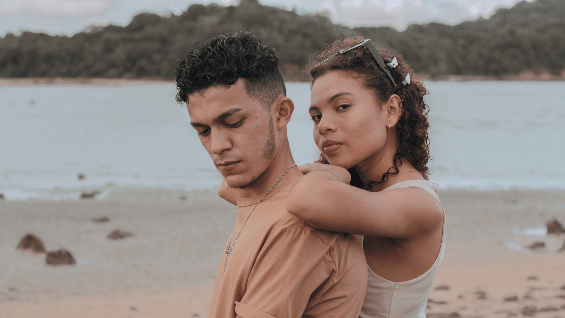 Foto de casal de namorados na praia, o homem está de costas para a mulher e olhando para baixo e ela está abraçando suas costas e olhando para frente