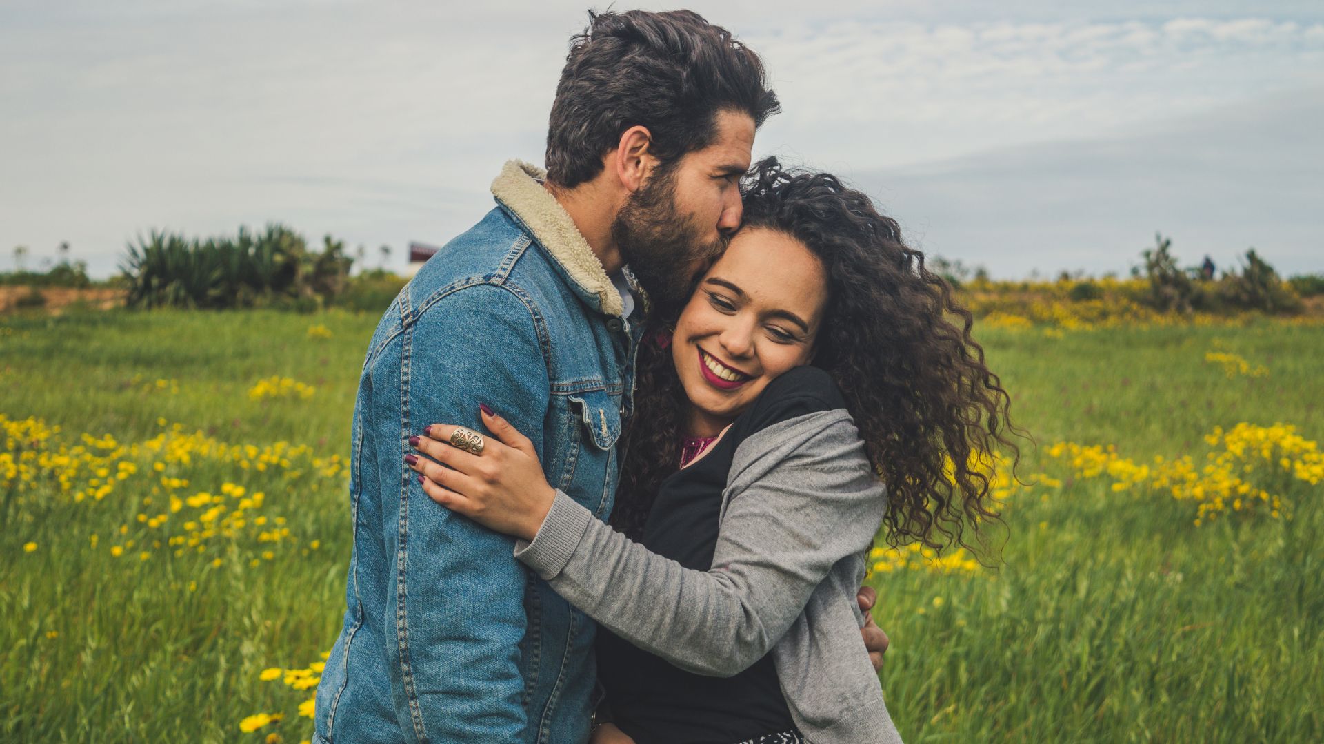 Imagem de um homem beijando o rosto de uma mulher que está sorrindo, em um campo com flores amarelas ao fundo