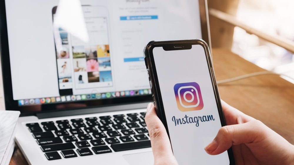 Pessoa segurando celular com o instagram aberto e o computador ao fundo com a rede social também aberta