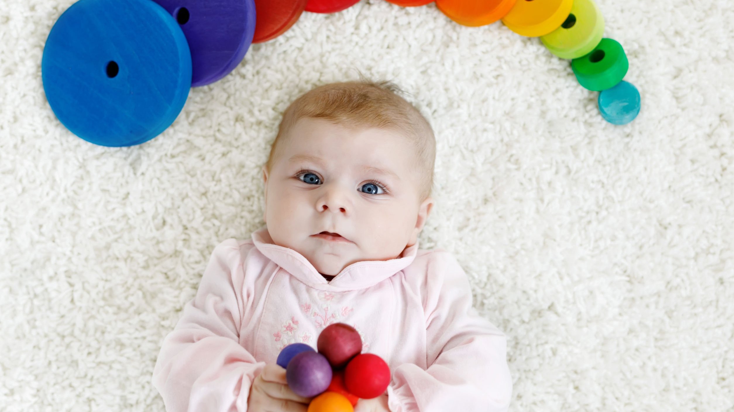 Bebê branco de olhos azuis deitado num tapete branco com brinquedos coloridos ao redor.