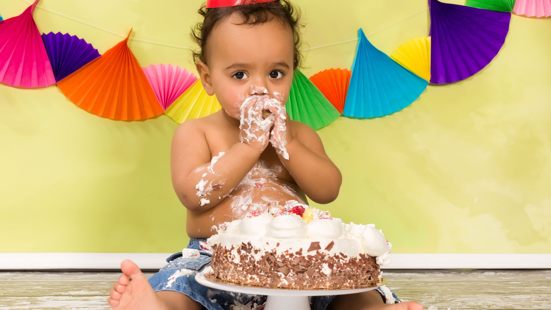 Imagem de um bebê comendo bolo de aniversário com a mão, atrás dele tem enfeites de festa.
