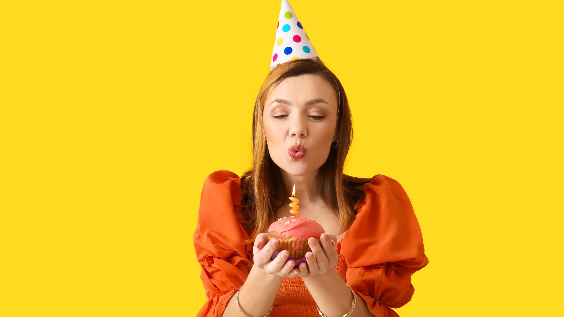 Imagem de uma mulher com roupa laranja e chapéu de aniversário colorido segurando um cupcake com uma vela em cima e assoprando a vela, em um fundo amarelo