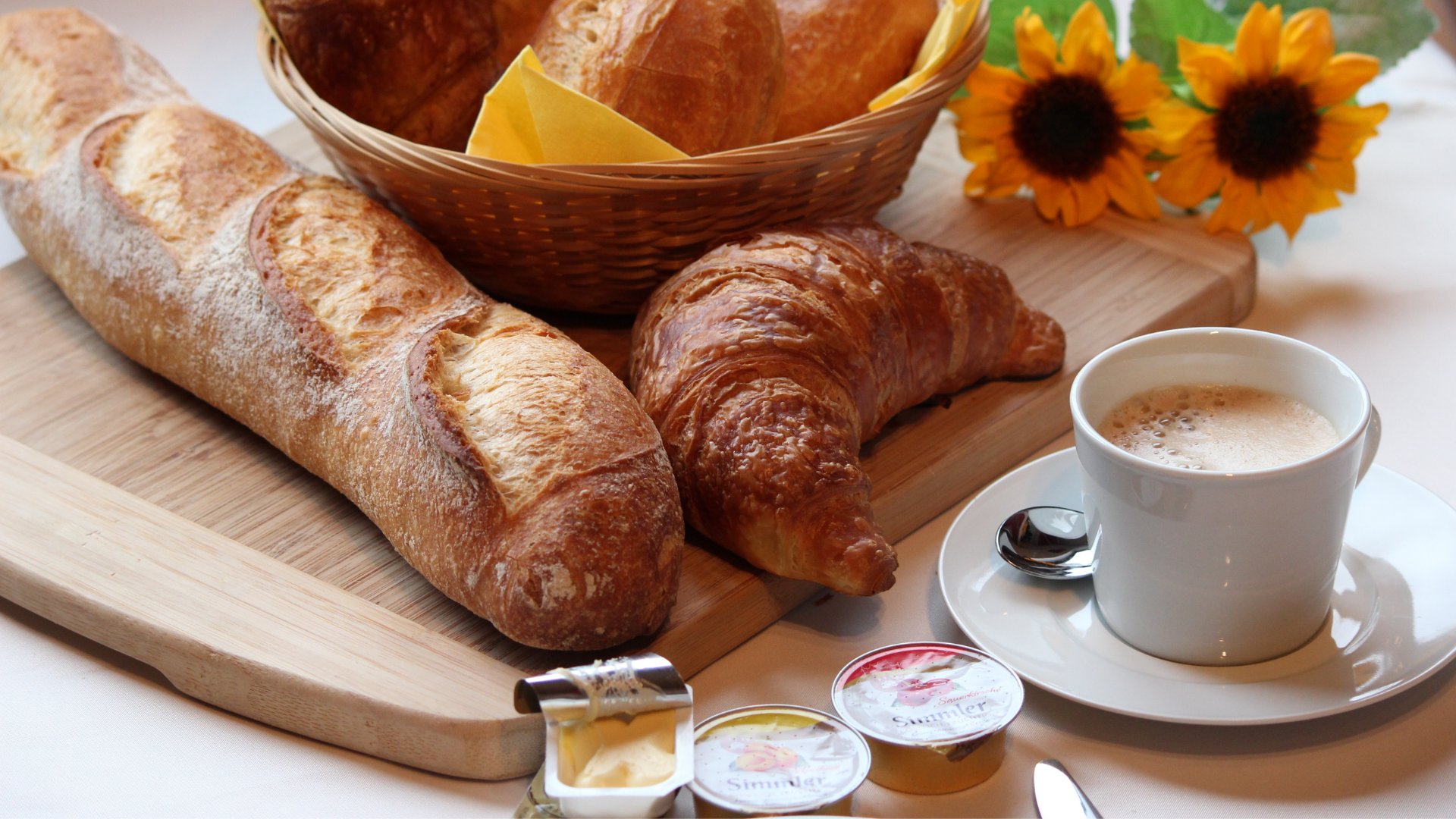 Maaci--335618 / Pixabay / Canva Caneca branca com café ao lado de pães e flores