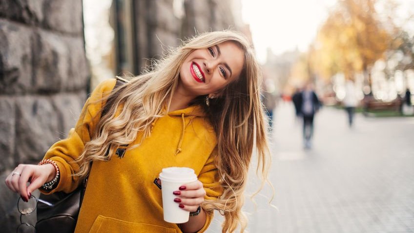 Jovem sorridente vestindo blusa amarela, enquanto segura um café na mão.