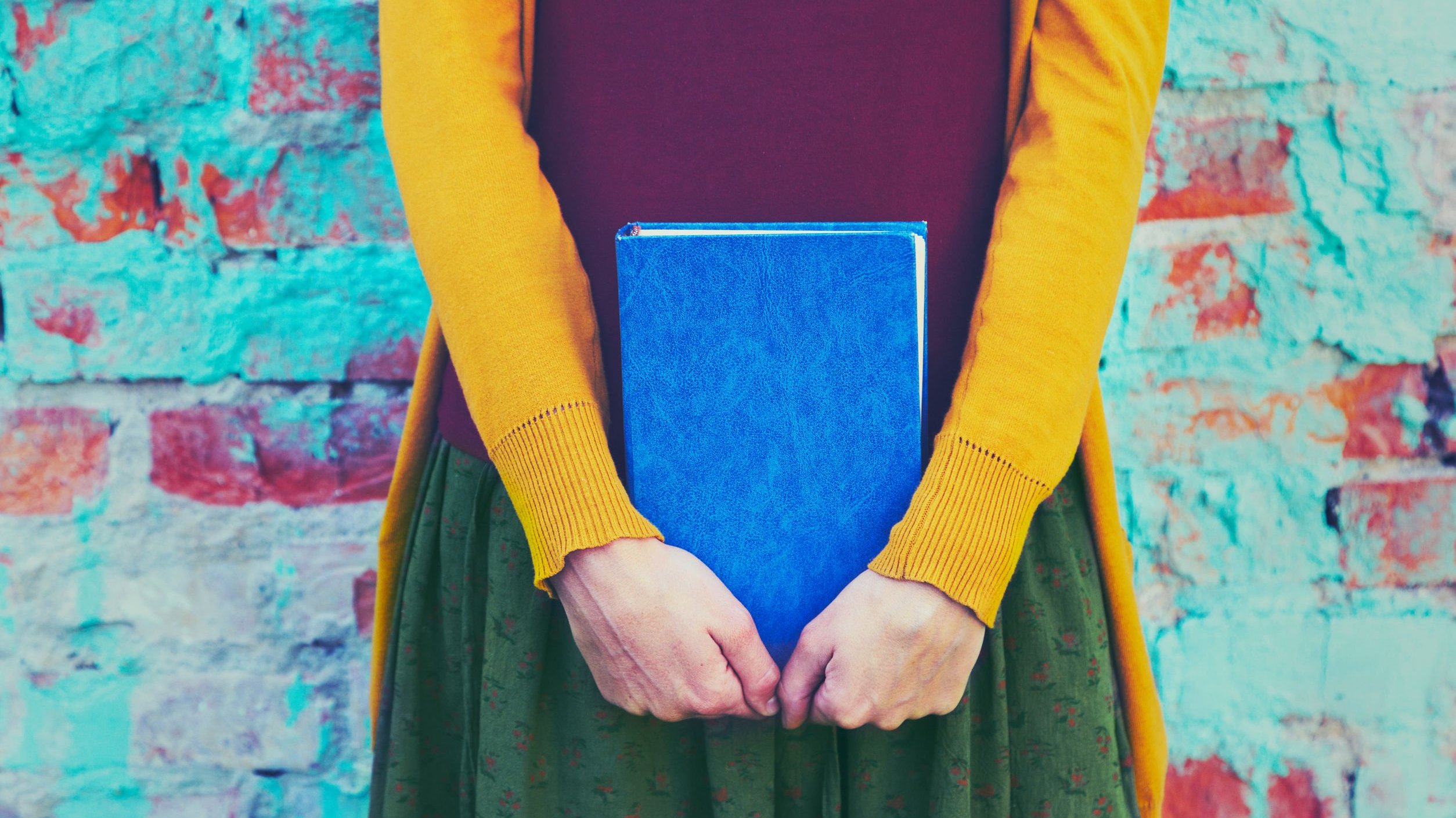 Mulher branca vestido roupa coloridas enquanto segura um livro azul.