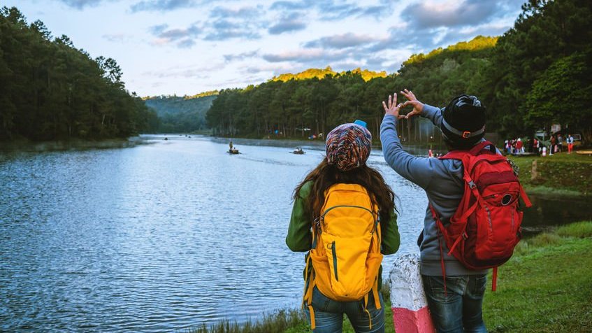 Casal de costas com mochilas de viagem e paisagem de lago ao fundo