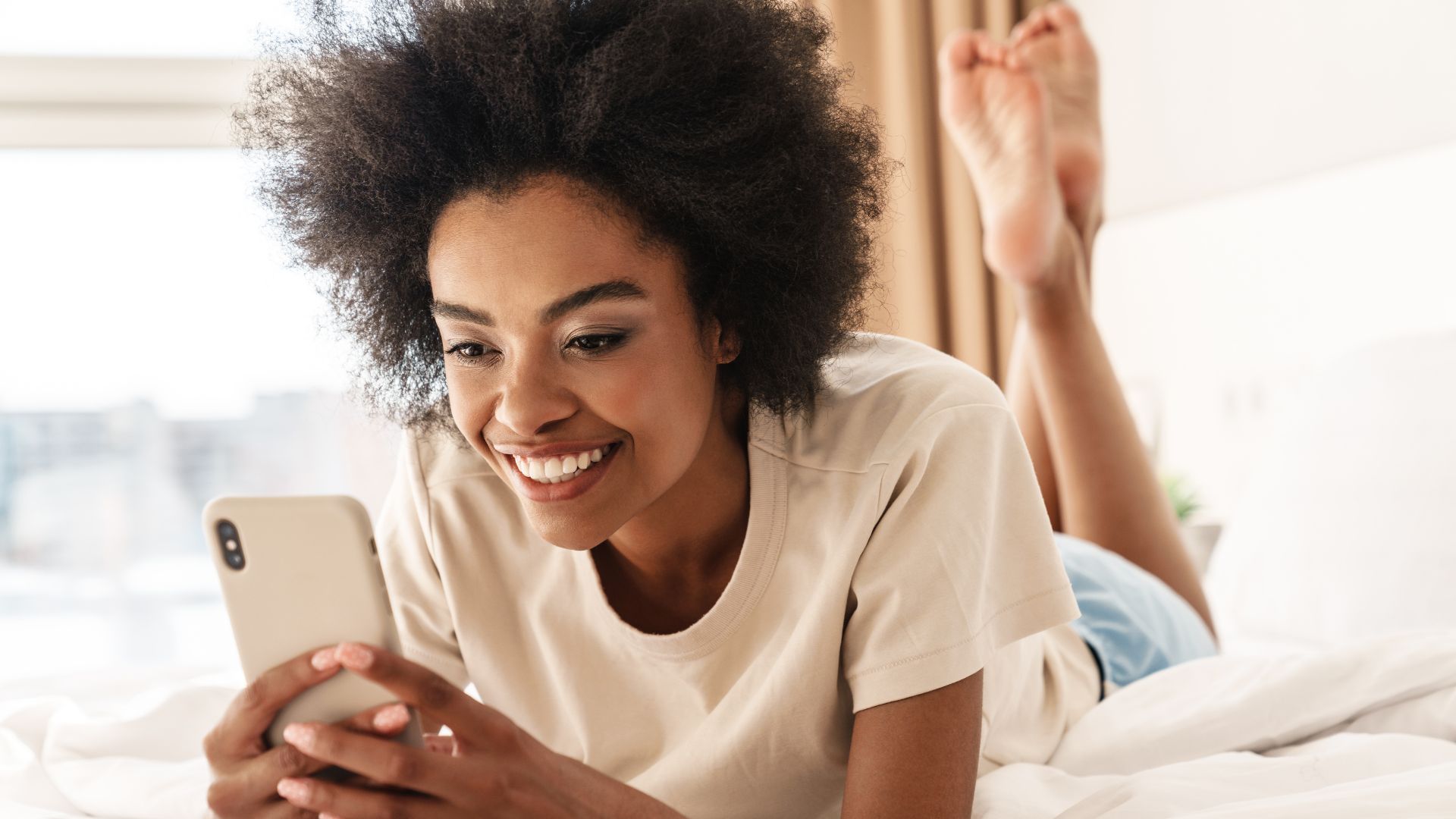 Imagem de uma mulher negra sorrindo enquanto vê o celular e na cama de barriga pra baixo.