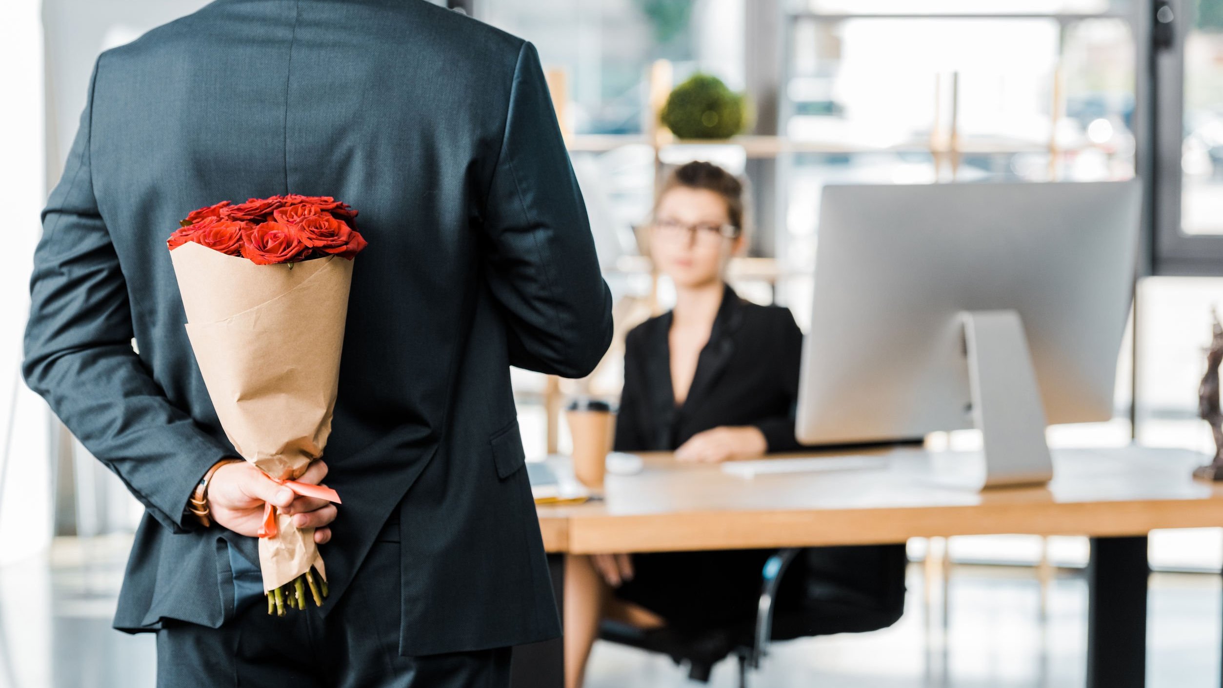 Homem segurando buquê de rosas atrás do corpo e mulher sentada atrás de mesa de escritório.