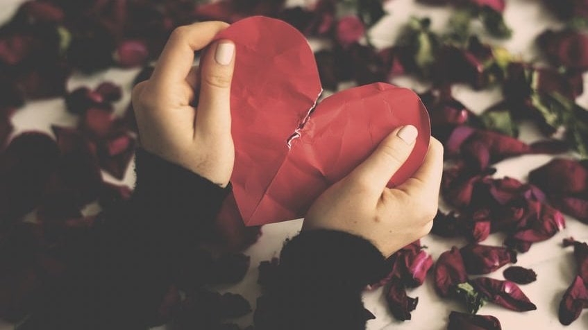 Recorte de uma mão segurando um coração partido com pétalas de rosas no fundo.