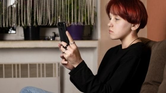 Adolescente sentada olhando fixamente para o celular que segura em suas mãos.
