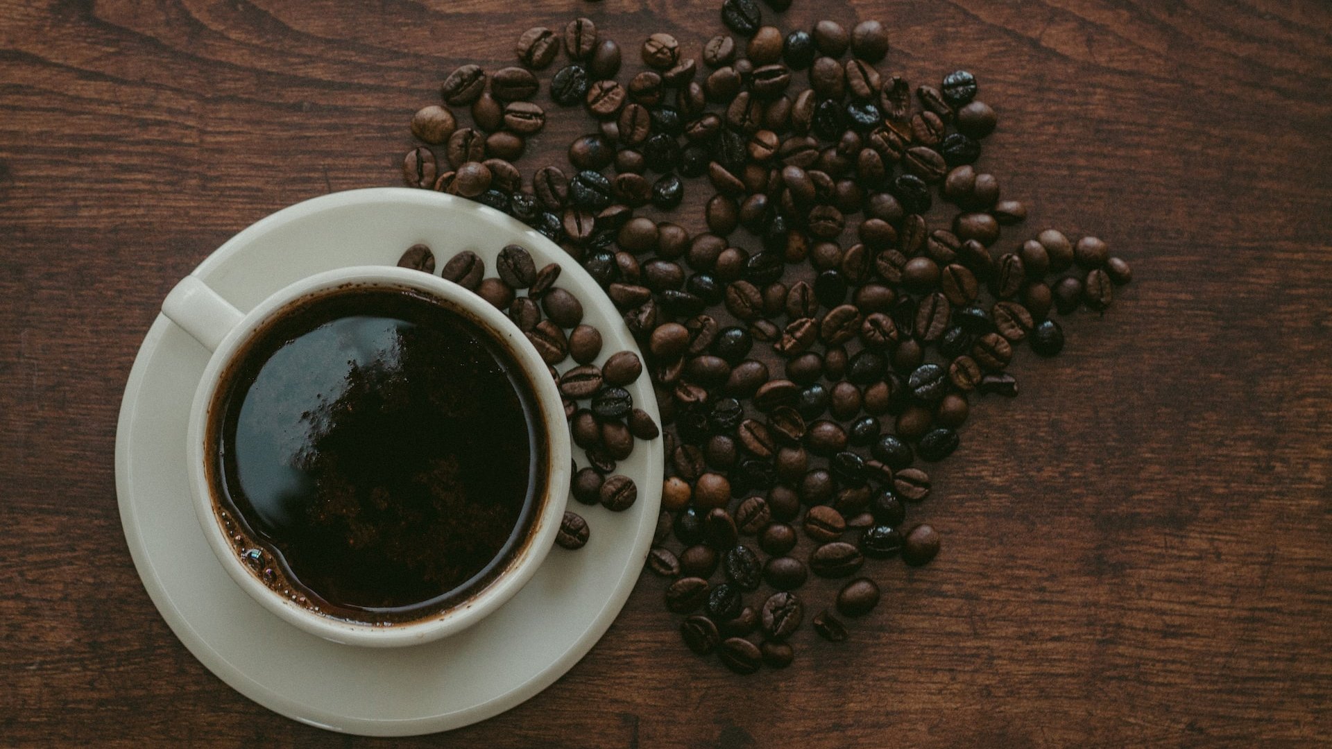 Xícara de café vista de cima. Ao seu lado, vários grãos de café reunidos sobre a superfície de madeira.