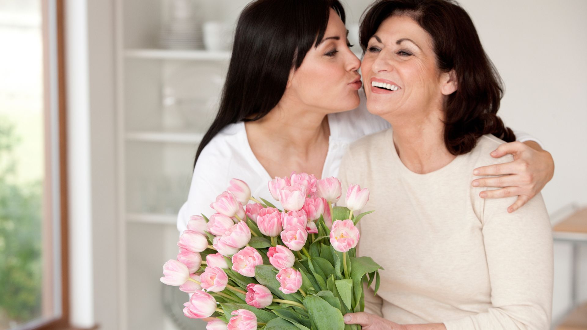 Imagem de uma filha abraçando a sua mãe e entregando um ramalhete de flores na cor rosa. Ambas usam blusa de frio nas cores bege e branco.