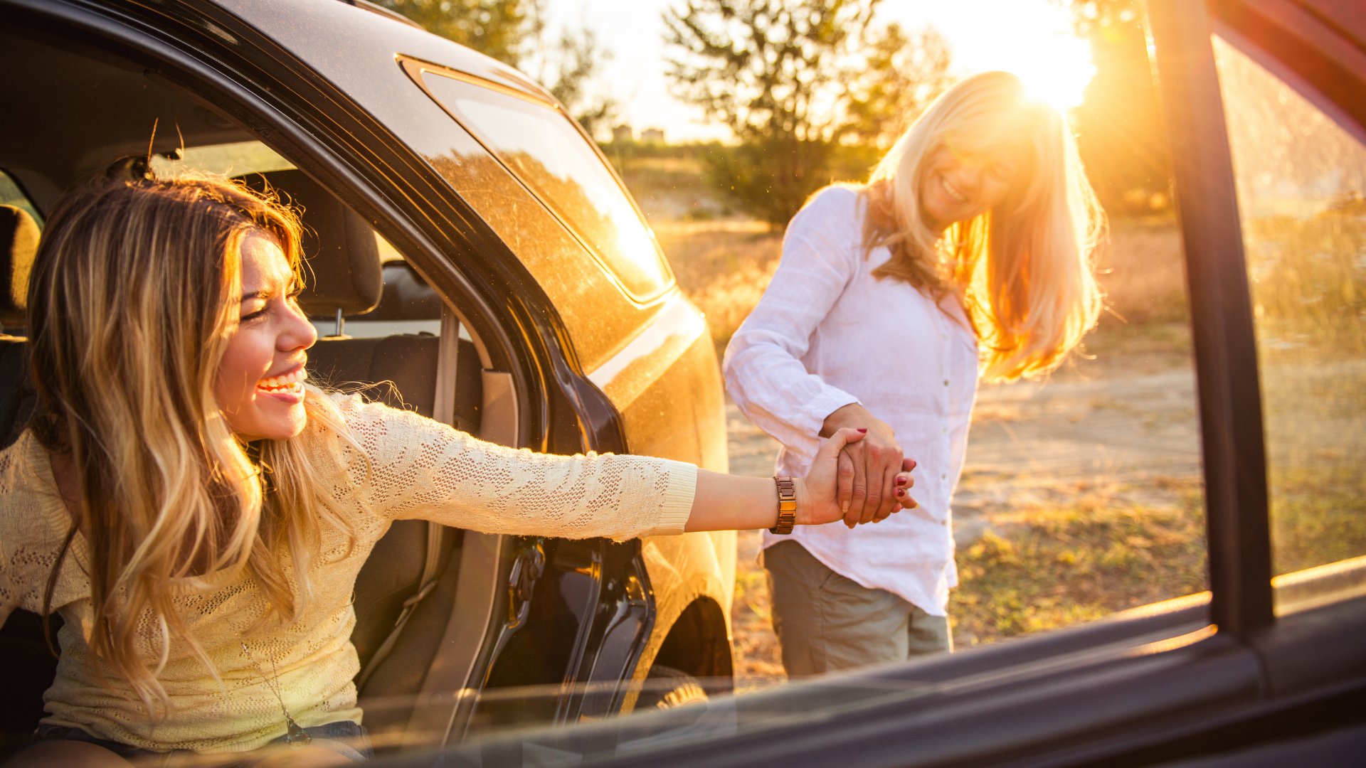 Mãe de mãos dadas com a filha que está dentro do carro enquanto o sol se põe