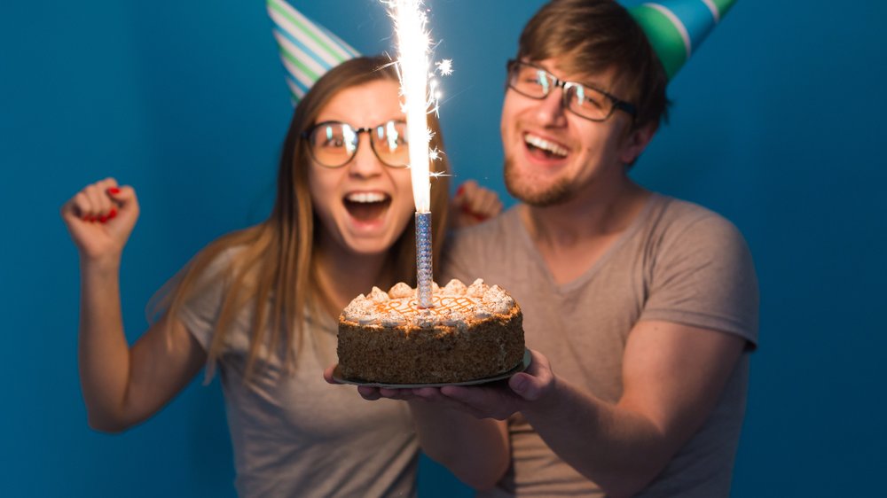 Tatiana Chekryzhova / Shutterstock.com Homem e mulher sorrindo para o bolo com uma vela soltando faíscas