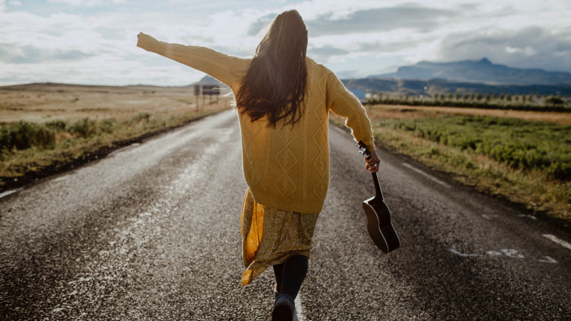 Imagem de uma mulher de costas, vestindo um vestido e uma blusa de lã amarela. Ela está andando por uma estrada e segurando um violão em uma das mãos.