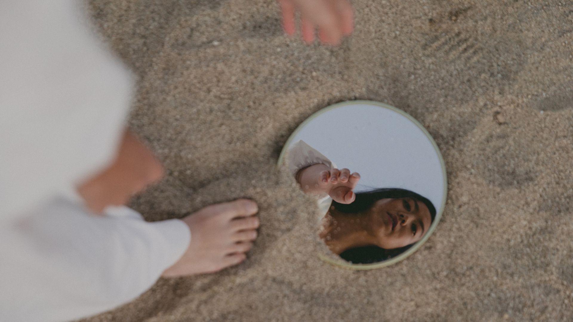 Imagem do reflexo de uma mulher em um espelho que está no chão, da areia da praia.